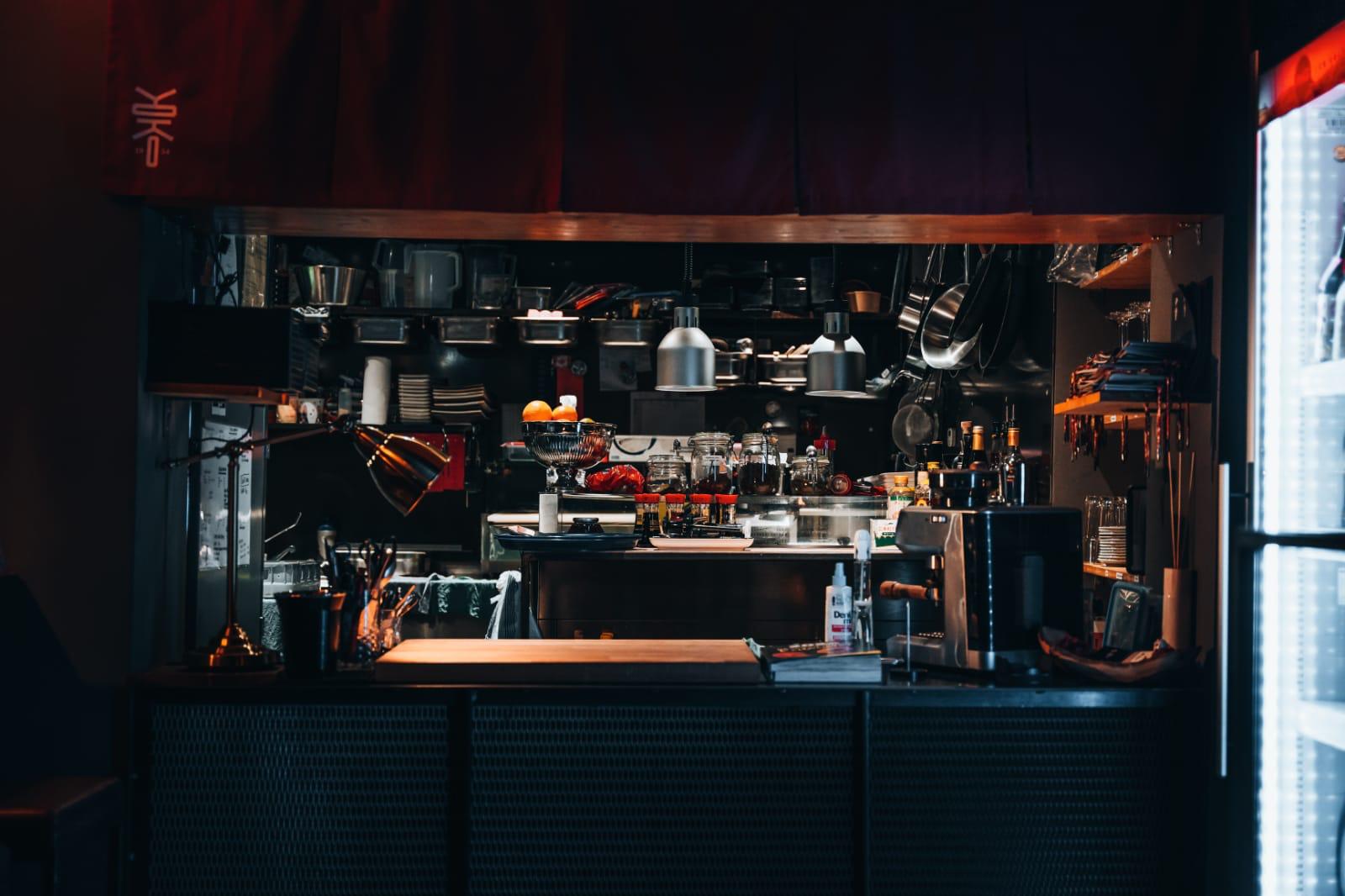 Blick in die offene Küche des Restaurants Yoko in Ulm mit Kochutensilien, Zutaten und warmem Licht, teilweise verdeckt von einem roten japanischen Vorhang mit Logo.