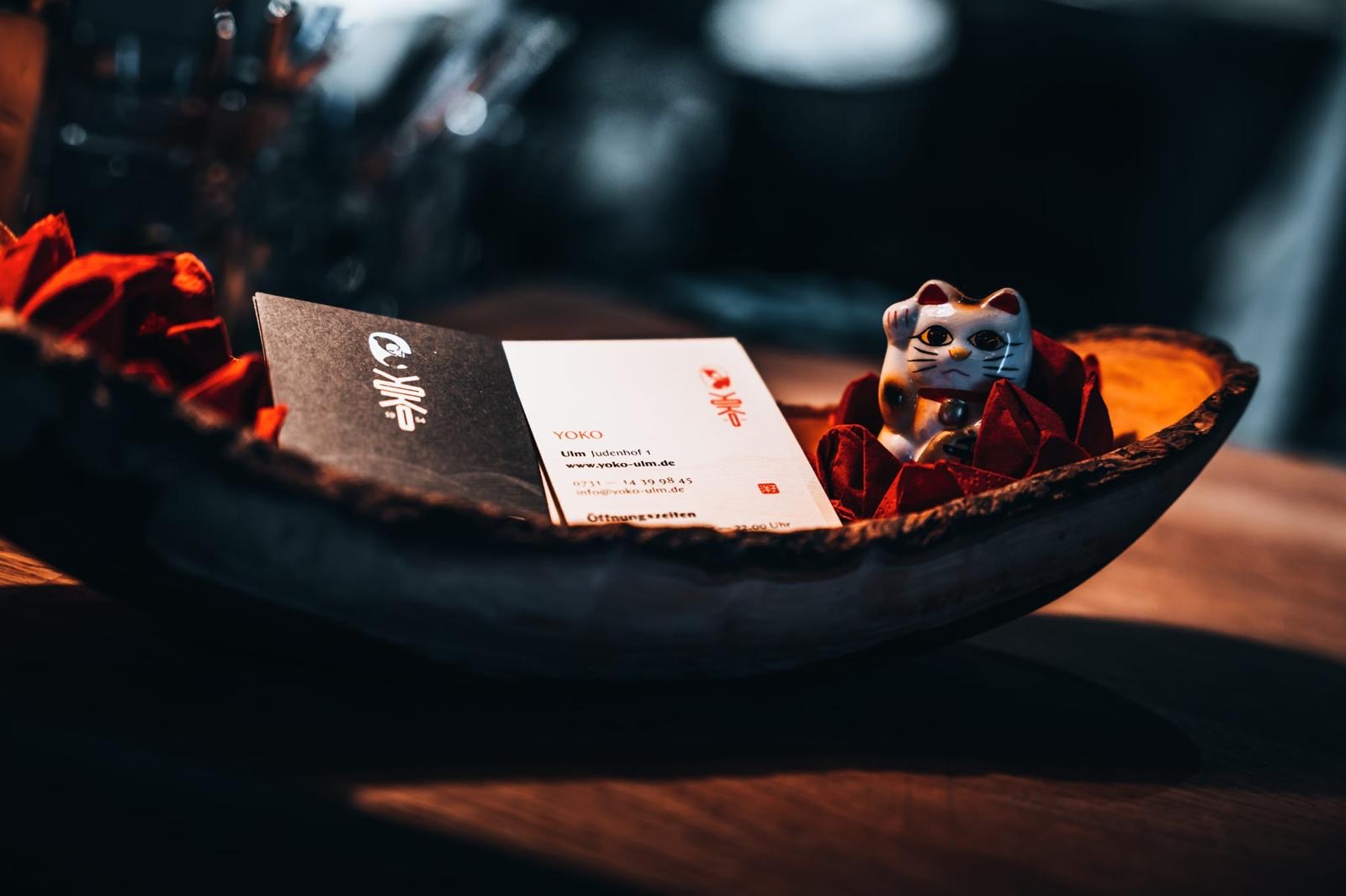 Nahaufnahme einer Schale mit roten Servietten, einer Visitenkarte des Restaurants Yoko in Ulm und einer kleinen winkenden Glückskatze.